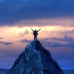 a person stands arms outstretched on the top of a mountain with a beautiful sunrise in the sky