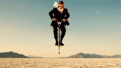 A man in business suit wearing old fashioned pilot's leather headgear, goggles and scarf bounces on a pogo stick in a dry, arid environment with nothing else around except distant hills in the background.