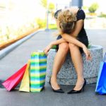 an exhausted shopper slumps on an outdoor seat with various coloured shopping bags either side of her.
