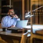 a man sits in unhappy contemplation staring at his computer on his desk in a home environment, propping his chin on his hand.