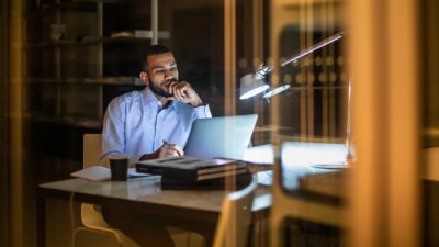 a man sits in unhappy contemplation staring at his computer on his desk in a home environment, propping his chin on his hand.