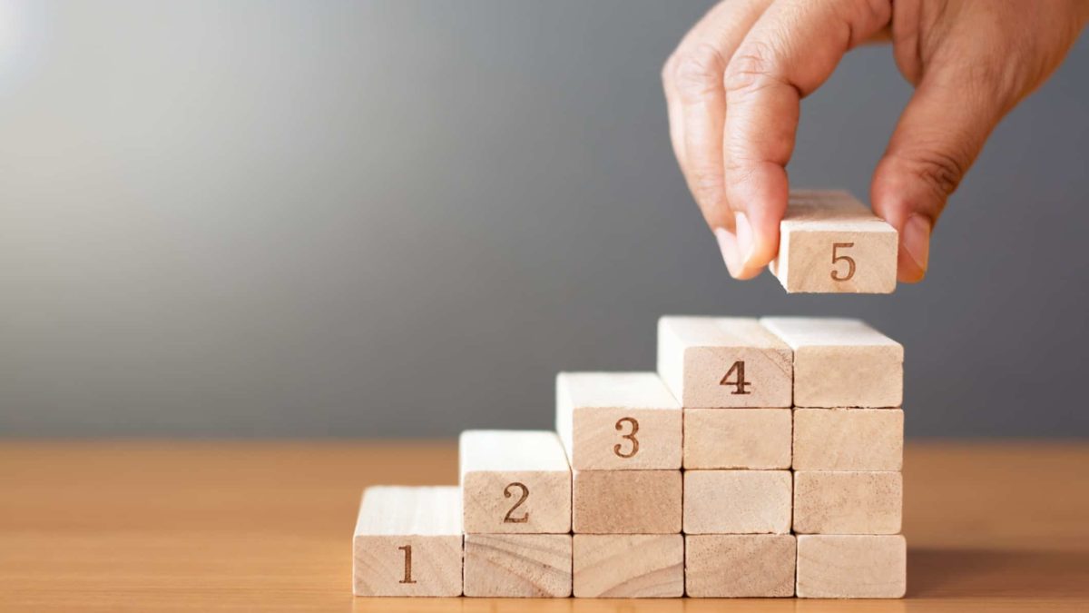 a hand places the number five on top of a pile of ascending wooden blocks, numbered 1 to 4 respectively. The number 5 pile is the tallest.