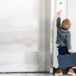A small child carrying a brief case tries to reach an elevator button outside closed elevator doors.