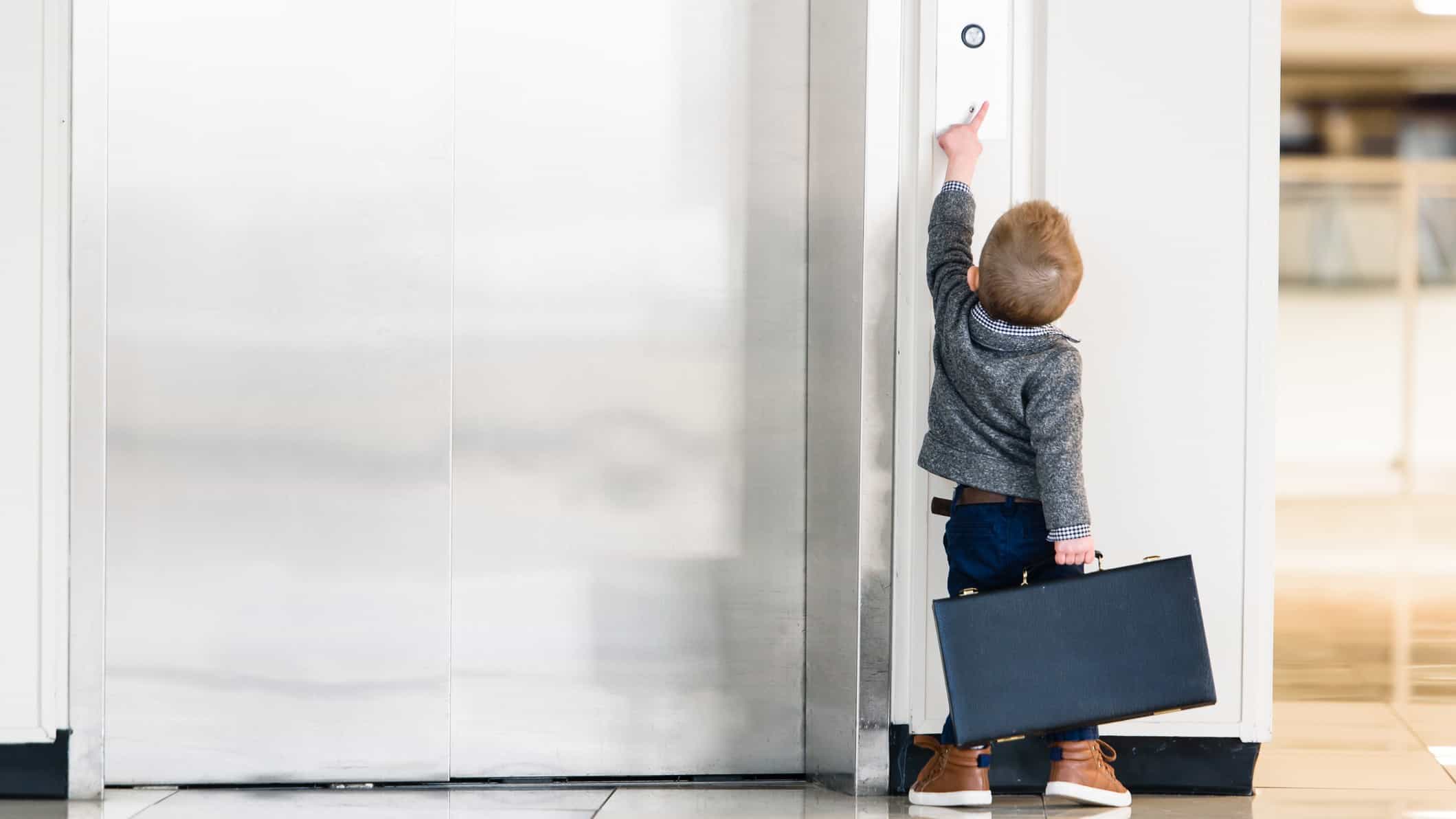 a small child carrying a brief case tries to reach an elevator button outside closed elevator doors.