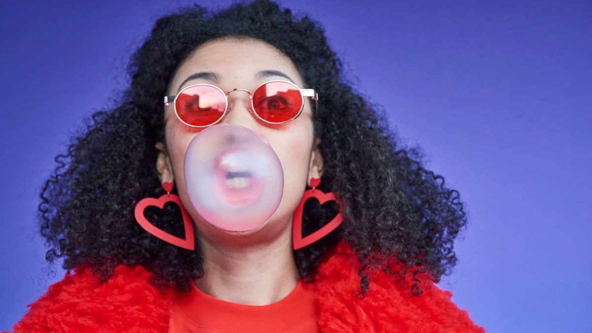 a woman wearing red blows a big bubble with bubblegum from her mouth.
