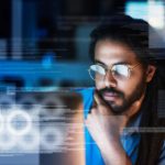 a man sits at a computer in deep thought with hand on chin in a darkened room as though it is late and night and he is working on cybersecurity issues.
