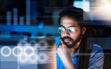 a man sits at a computer in deep thought with hand on chin in a darkened room as though it is late and night and he is working on cybersecurity issues.