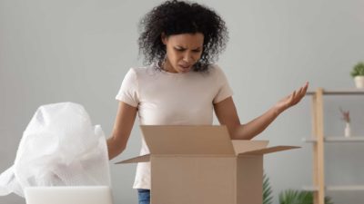 a woman looks disappointed with a package she has unpacked holding her arms up at a box with bubble wrap beside it.