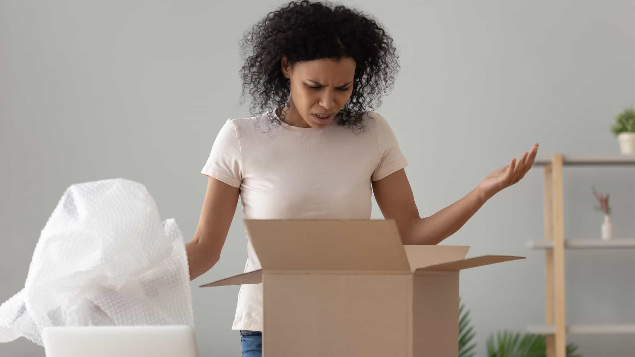 a woman looks disappointed with a package she has unpacked holding her arms up at a box with bubble wrap beside it.
