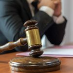 A gavel is placed on a stand on a desk with a legal representative wearing a suit in the background.