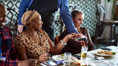a woman uses a card to pay at a restaurant, with the waiter leaning into the table where there is food and drink after a meal.