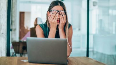 A woman sits with her hands covering her eyes while lifting her spectacles sitting at a computer on a desk in an office setting.