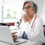 A doctor in a white coat sits at her computer with finger on mouth thinking about something in her office with medical equipment in the background.