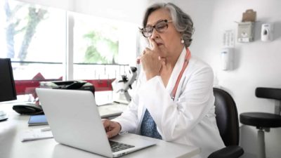 A doctor in a white coat sits at her computer with finger on mouth thinking about something in her office with medical equipment in the background.