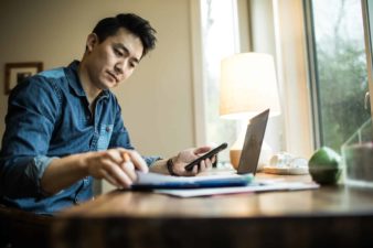 A blockchain investor sits at his desk with a laptop computer open and a phone checking information from a booklet in a home office setting.