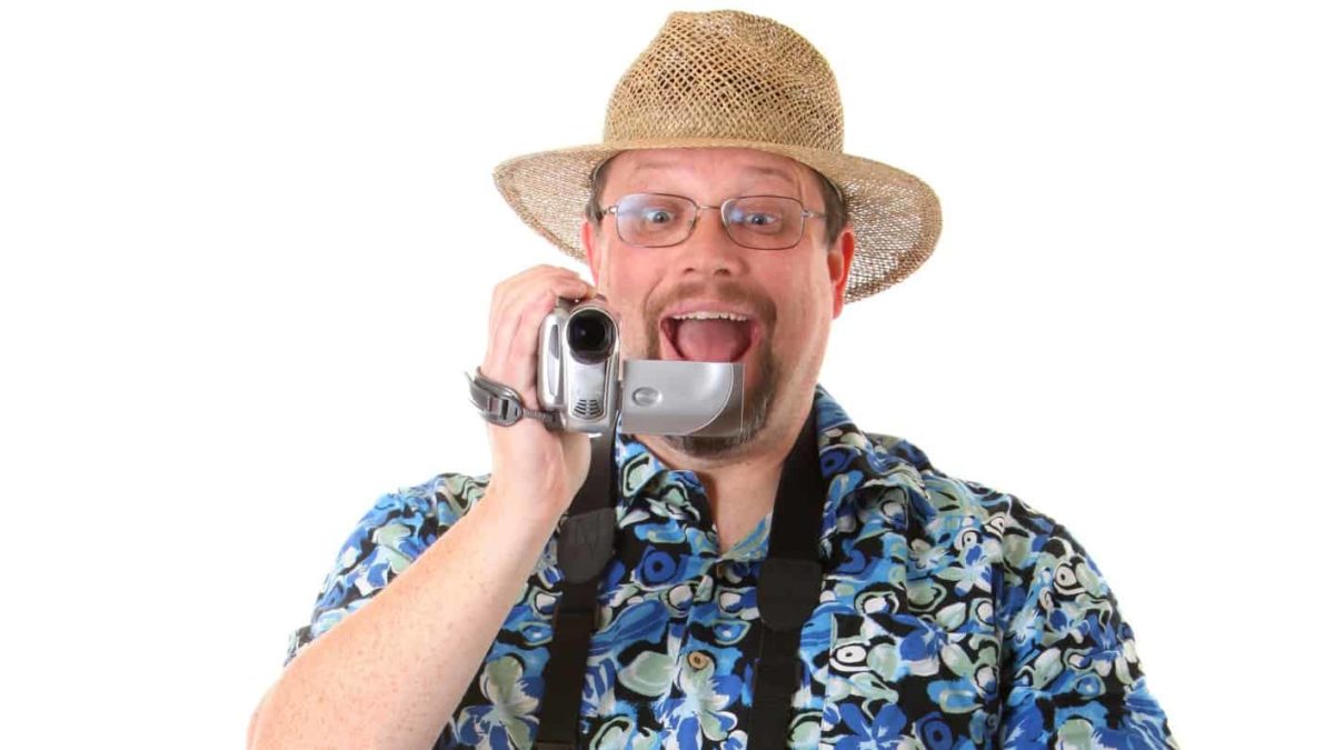 a tourist wearing a bright patterned shirt, straw hat and with a video camera has an excited look on his face.