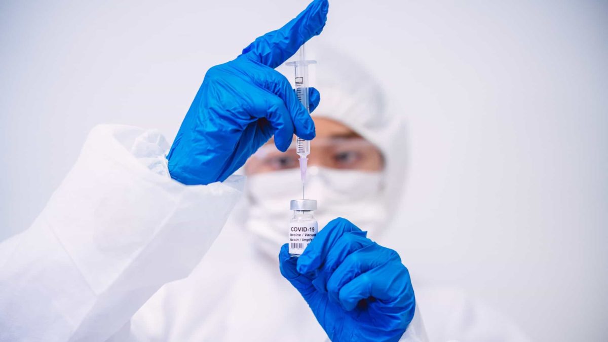 a medical researcher holds a vial of vaccine with a needle inserted in the top watching intently behind a mask and goggles and protective medical clothing.