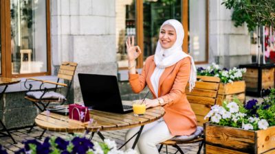 a woman sitting at a computer making an ok symbol with her hand, making a circle with her thumb and forefinger and holding the other three fingers up straight, as she sits in a colourful courtyard setting.