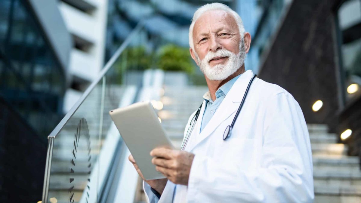 a doctor in white coat and stethoscope stands in front of a building holding an electronic device in his hands.