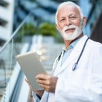 a doctor in white coat and stethoscope stands in front of a building holding an electronic device in his hands.