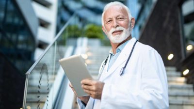 a doctor in white coat and stethoscope stands in front of a building holding an electronic device in his hands.