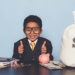 a happy child dressed in full business suit gives the thumbs up sign while sitting at a desk featuring a piggy bank and a sack of money with a dollar sign on it.