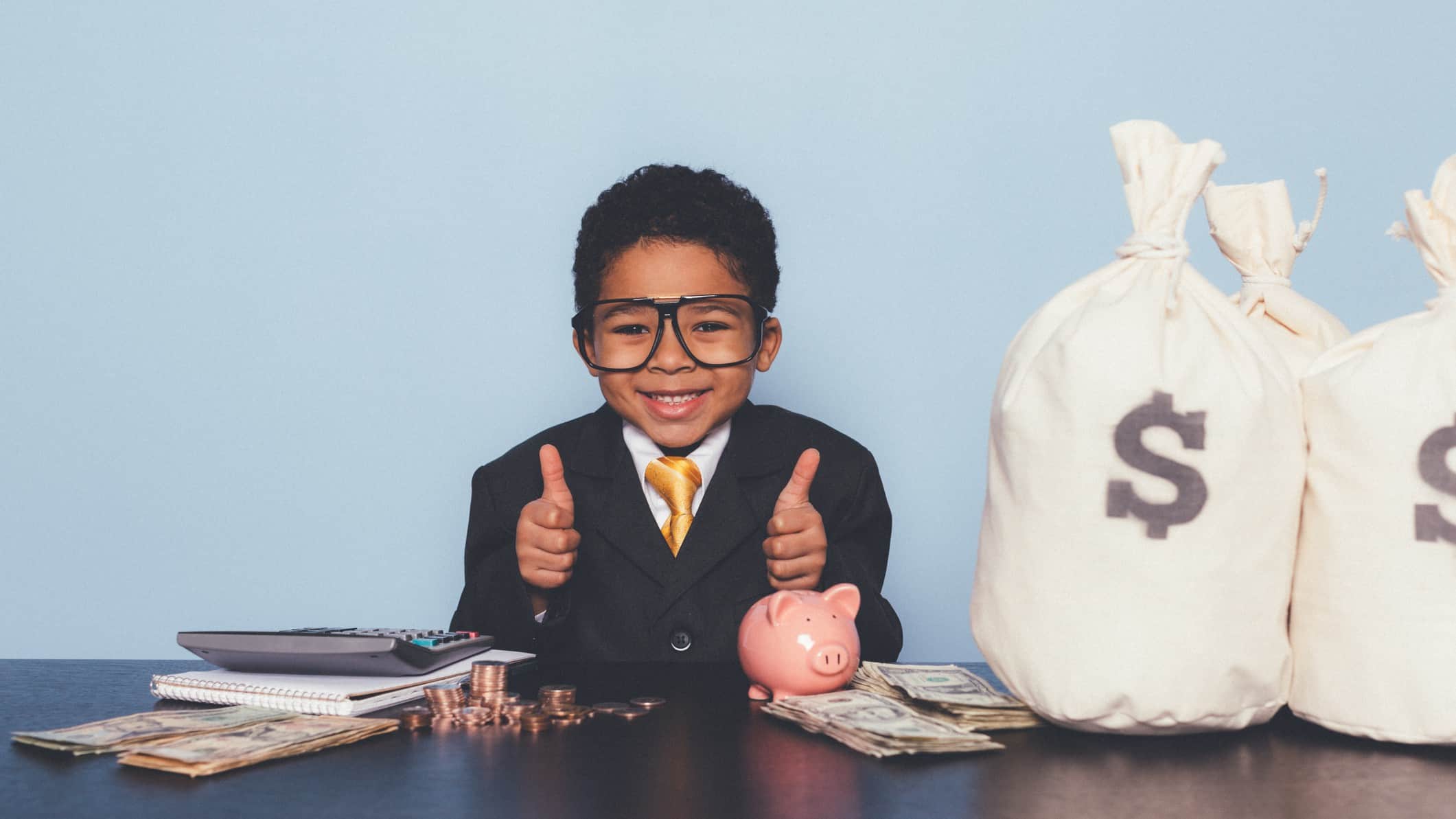 a happy child dressed in full business suit gives the thumbs up sign while sitting at a desk featuring a piggy bank and a sack of money with a dollar sign on it.