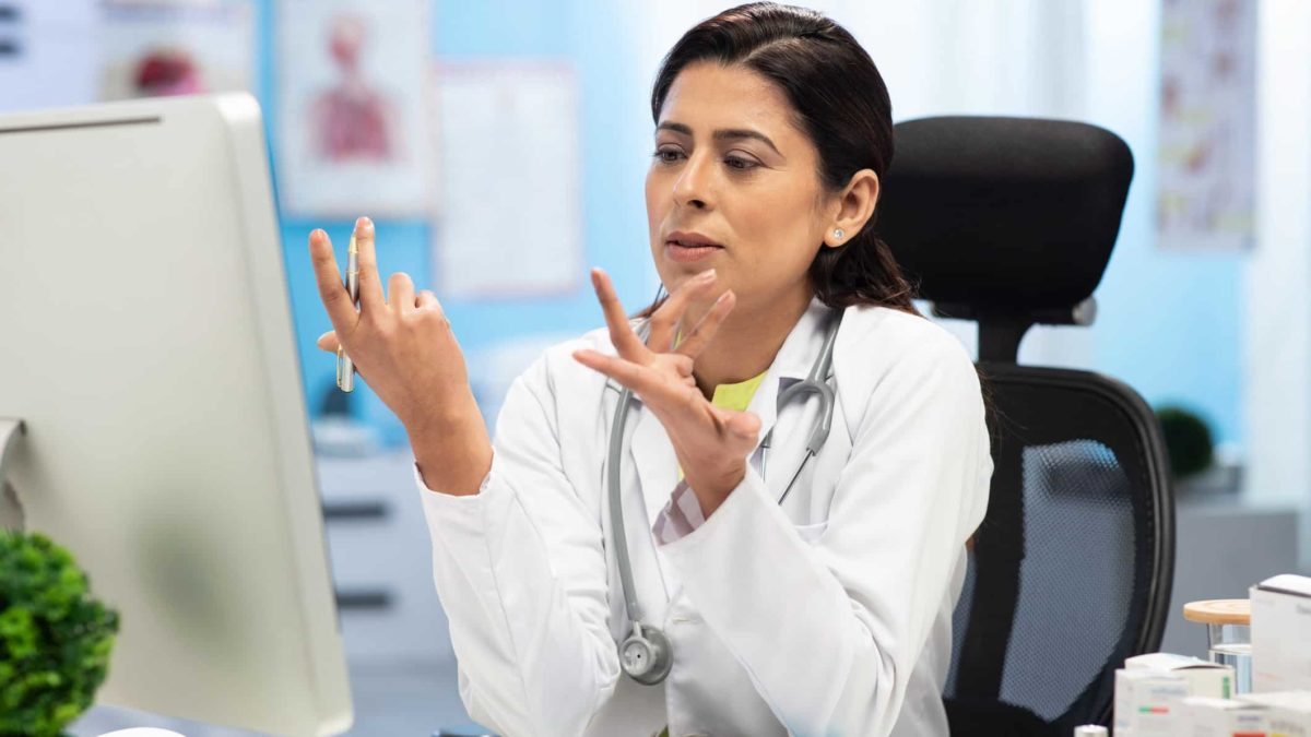 A doctor in a white coat with a stethoscope around her neck holds her hands upwards as if to ask 'why' as she sits at her desk and looks at her computer.