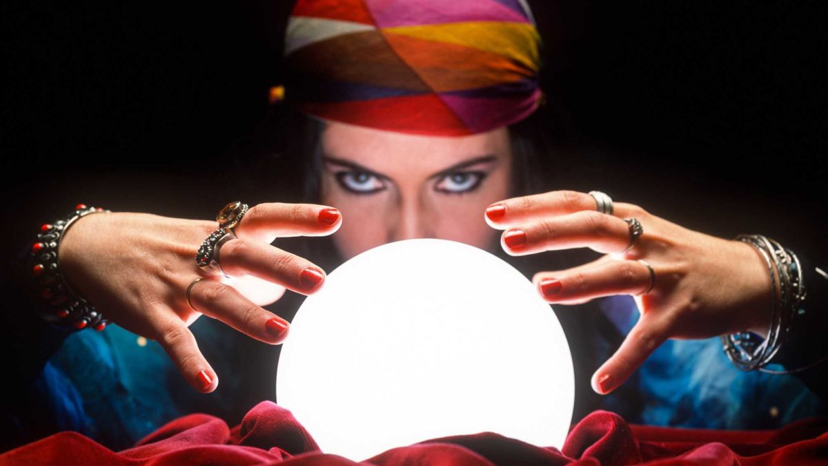 a woman with a colourful head scarf peeers over a brightly lit crystal ball casting her hands around it as if to predict the future.