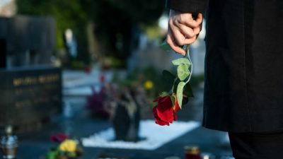 a hand of a person wearing a black coat holds a single red rose with a grave site and gravestone in the background.