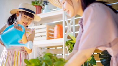 a young girl waters a plant with a watering can while her mother is tending to a pot plant in an indoor location.