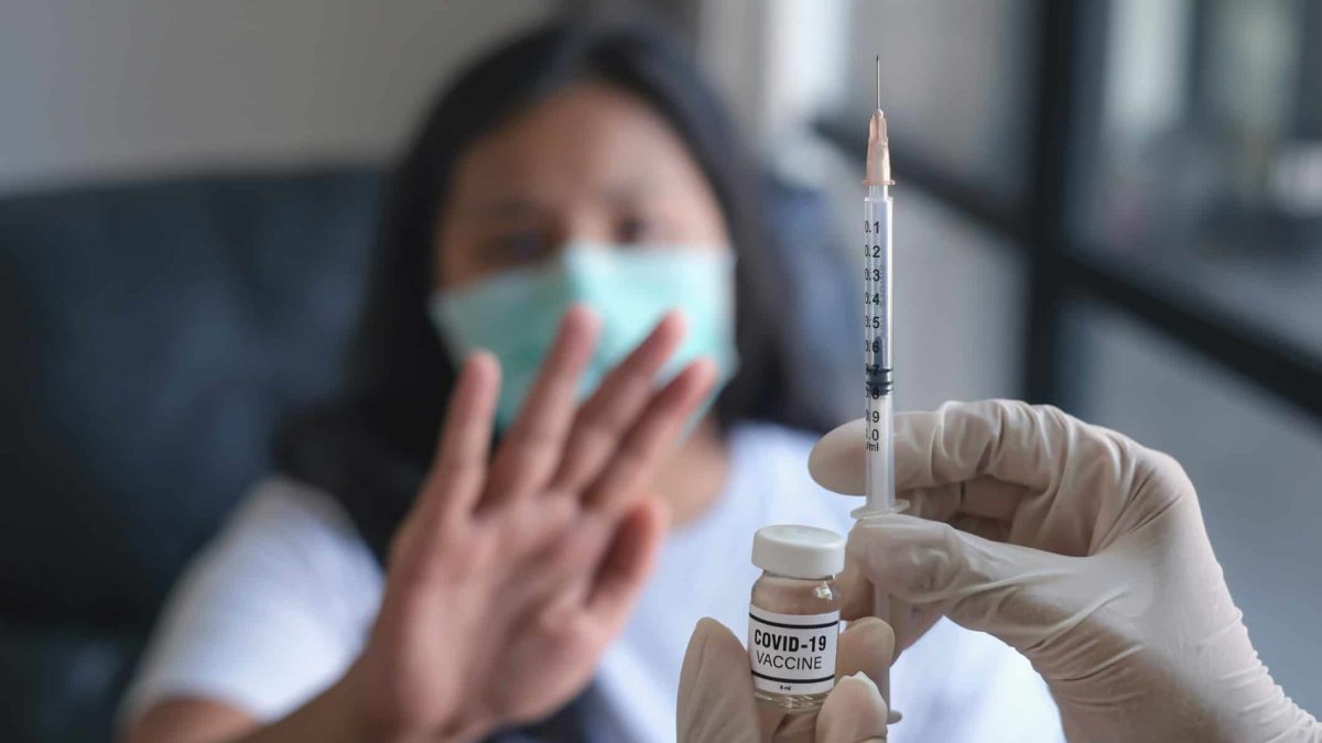 a woman in a mask holds up a hand to stop a COVID-19 vaccine being held by a medically gloved hand in the foreground.