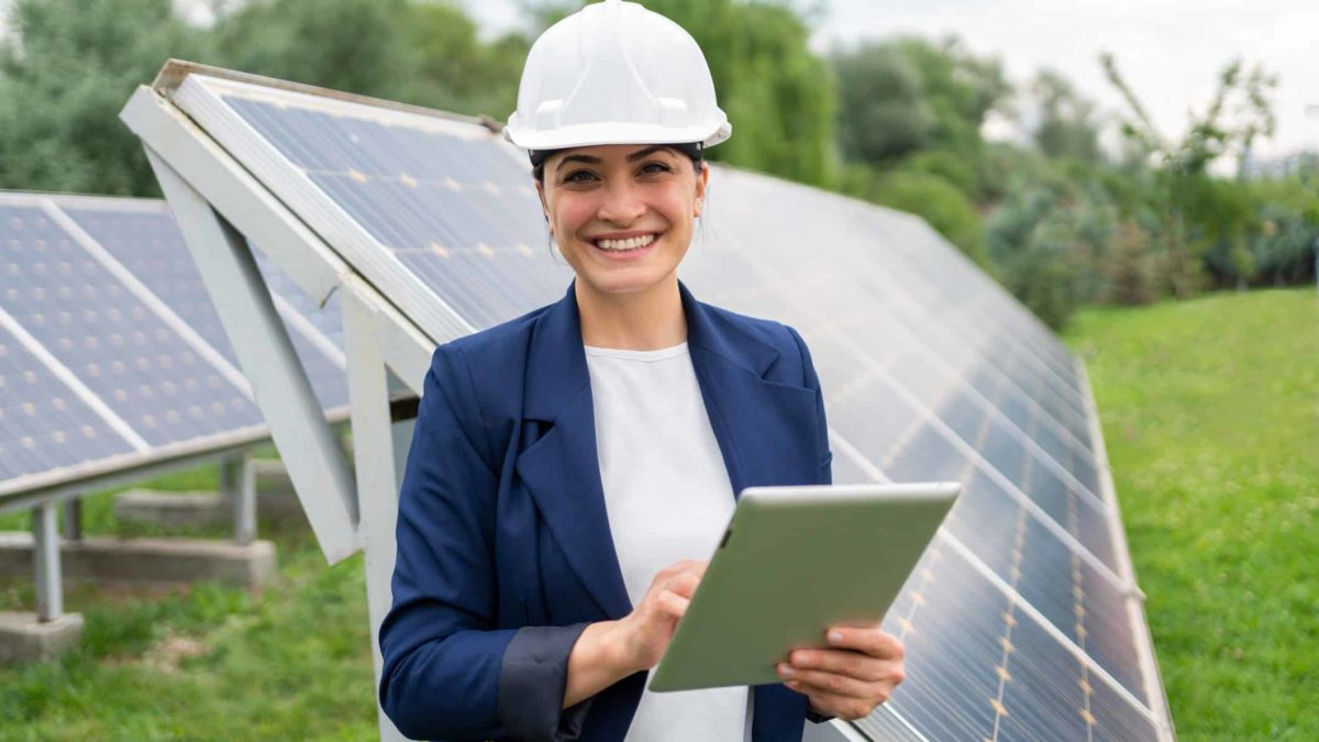 an engineer in hard hat stands amid solar panels, part of a solar farm, as she holds a tablet in her hand and smiles.