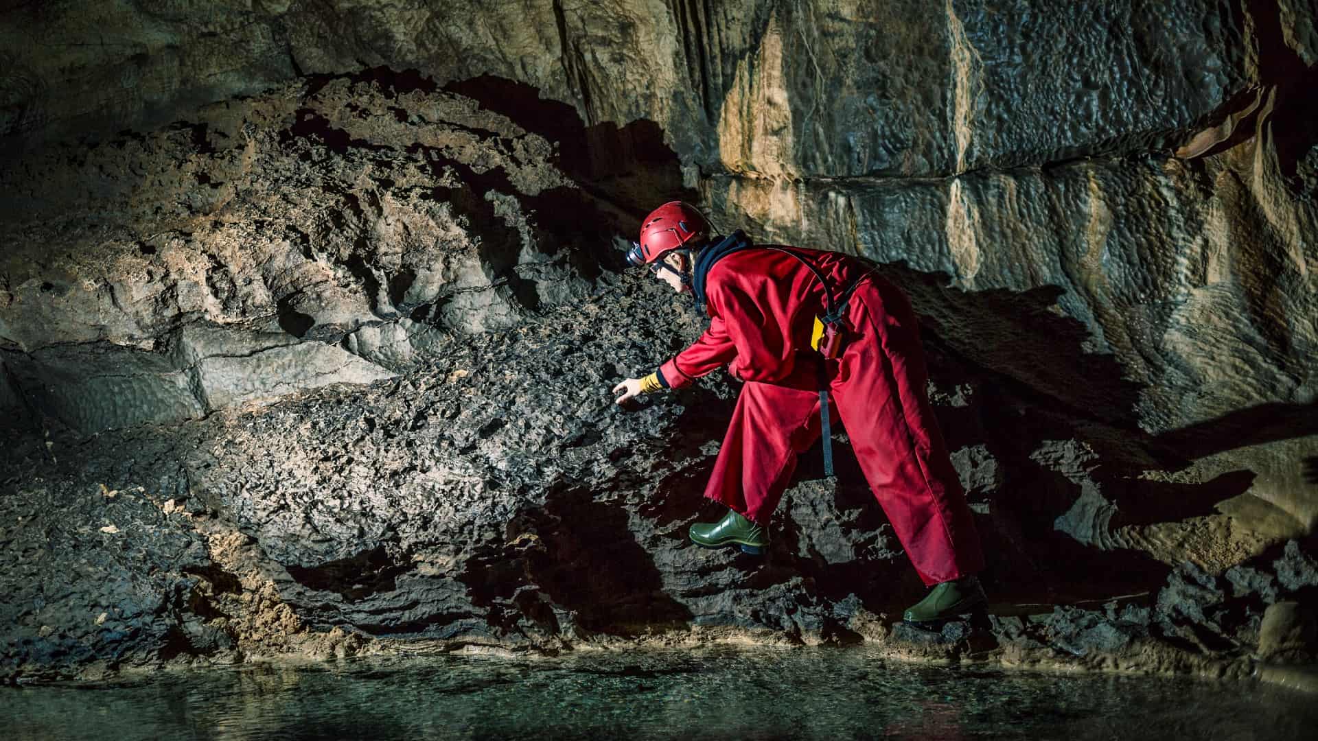 a geologist or mine worker looks closely at a rock formation in a darkened cave with water on the ground, wearing a full protective suit and hard hat.