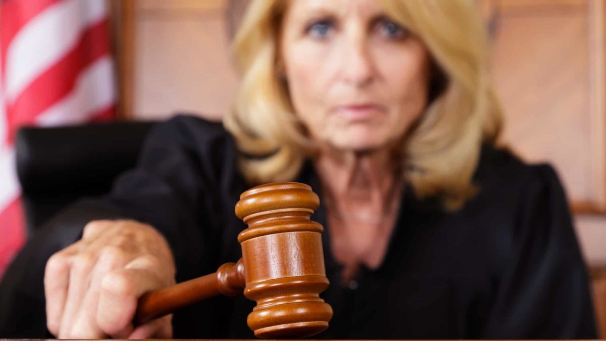 a stern judge slams a gavel onto her desk with the American flag visible in the background.