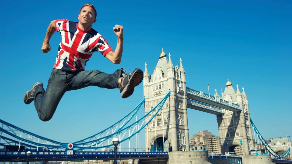 a man in a british union jack T shirt hurdles high into the air with london bridge visible in the background.
