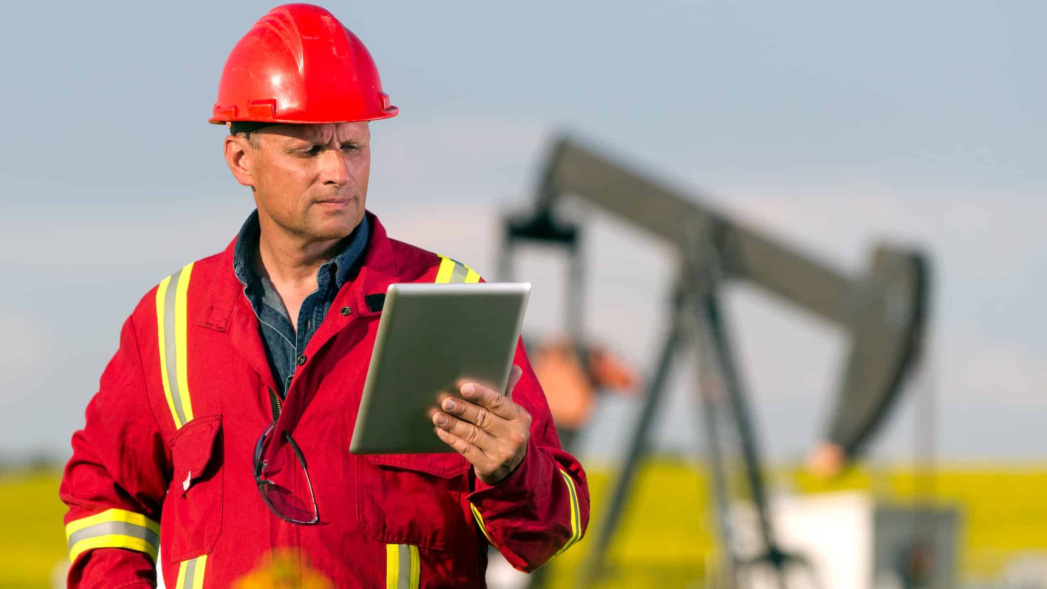 A miner in visibility gear and hard hat looks seriously at an iPad device in a field where oil mining equipment is visible in the background.