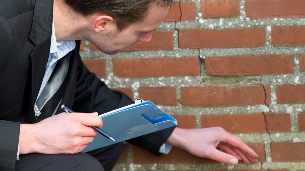 a person with clipboard and pen assesses a crack in a brick wall, perhaps caused by an earth tremor.