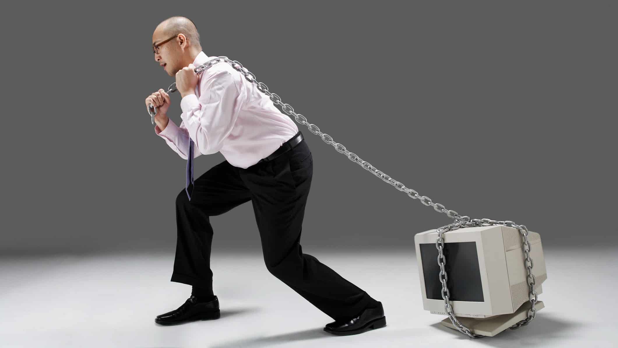 a man in a business shirt and trousers drags a chain wrapped around a computer as thought it is very heavy to move.