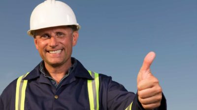 A miner in hardhat and high visibility clothing makes a thumbs up symbol against a blue sky.