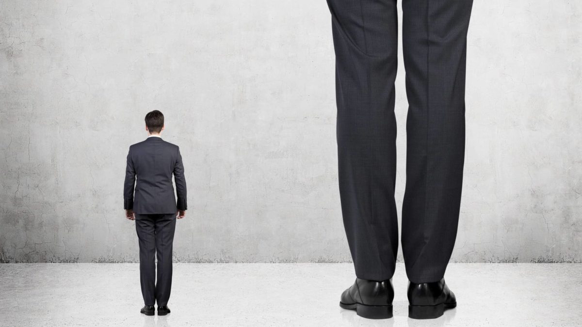 a small person in business attire stands next to a very tall person with only their legs in the image.