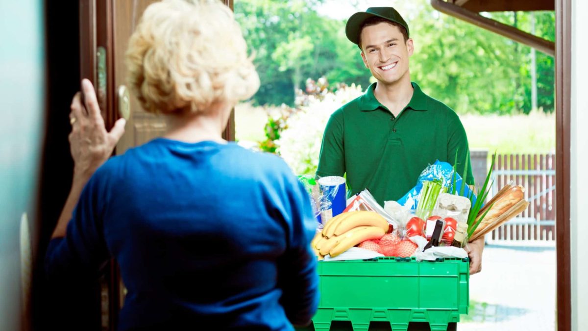 a grocery delivery worker stands at a front door with a large box of products while an older woman holds the door open to him.