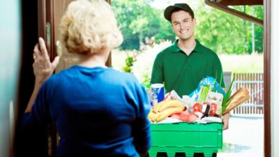 a grocery delivery worker stands at a front door with a large box of products while an older woman holds the door open to him.