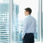 A man stares out of an office window onto a landscape of high rise office buildings in an urban landscape.