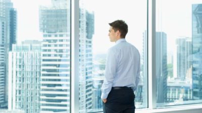 a man with hands in pockets and a serious look on his face stares out of an office window onto a landscape of highrise office buildings in an urban landscape