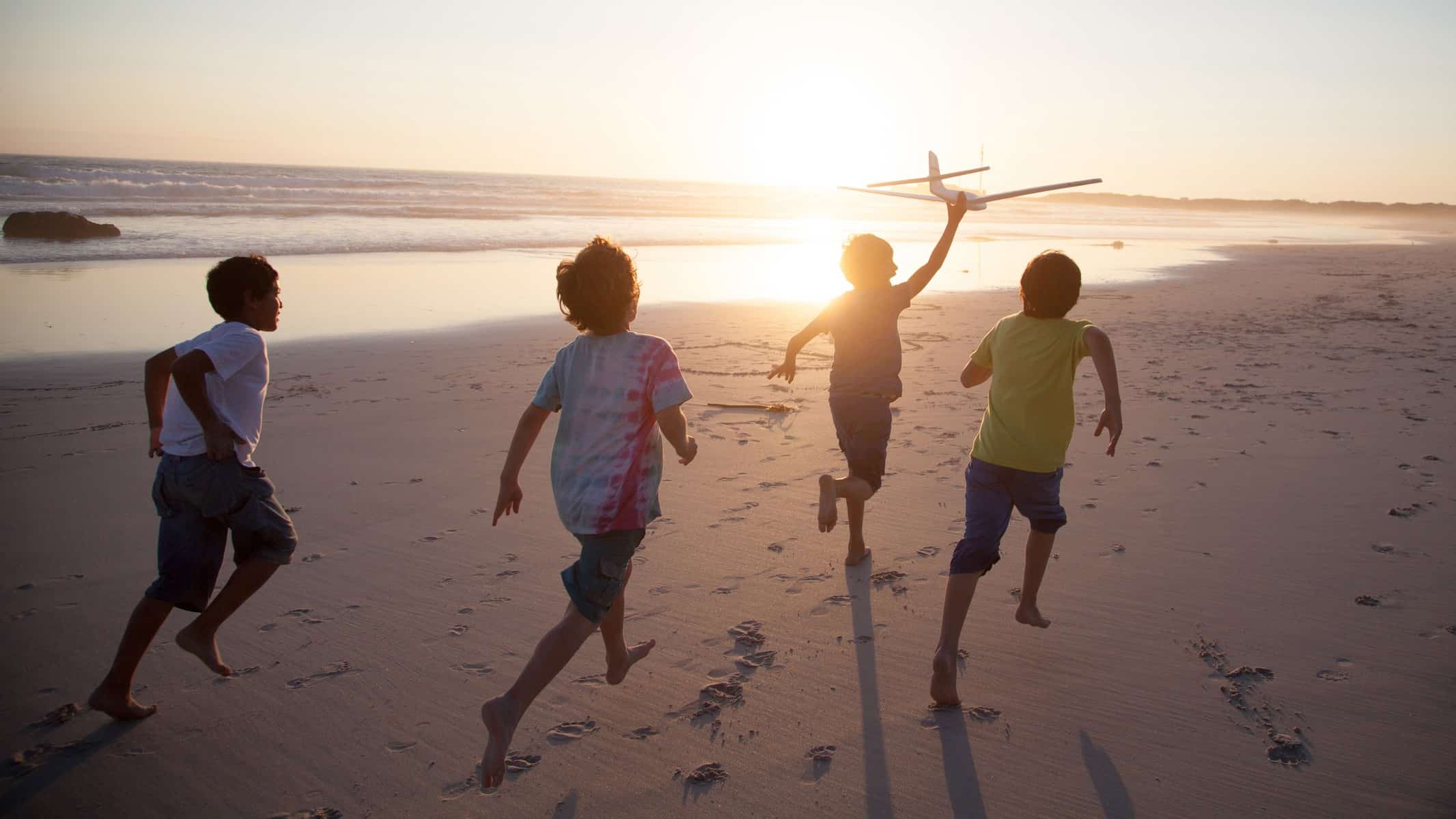 a group of four young boys run along a beach at sunset with the one in front holding aloft a toy aeroplane that is zooming through the air.