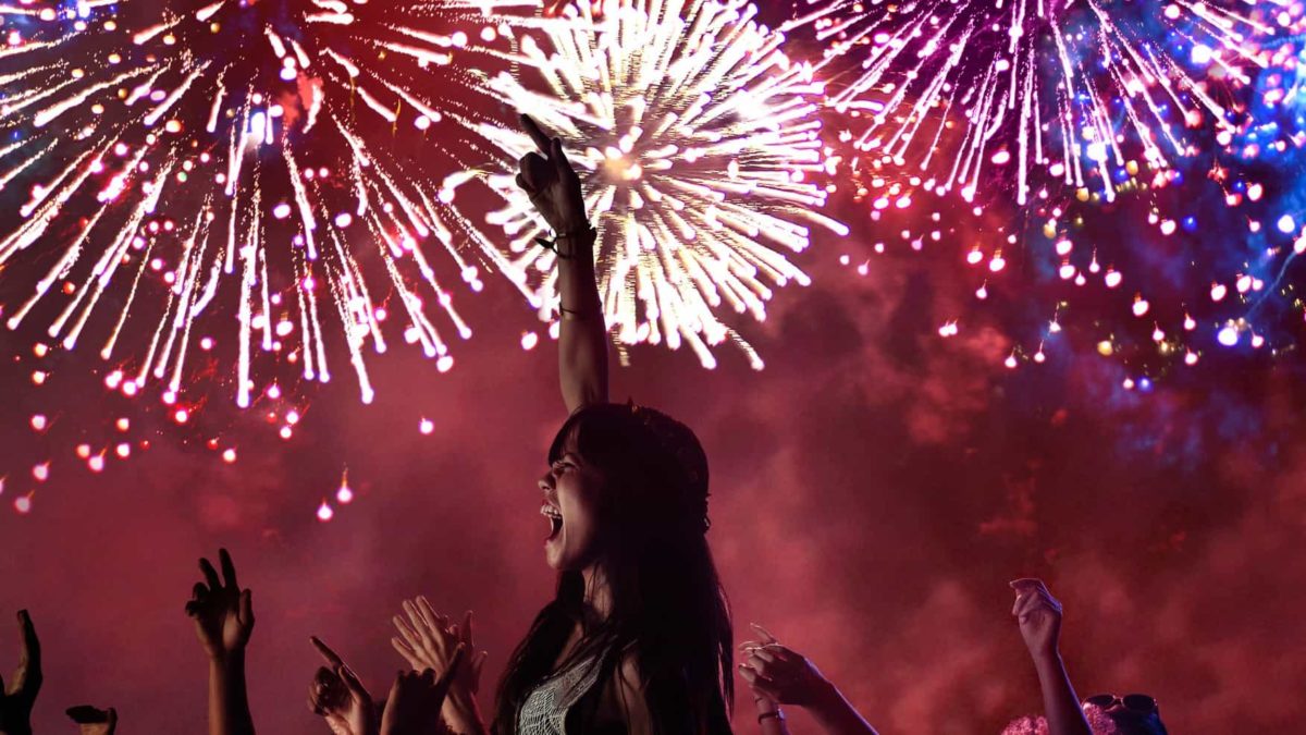 a young woman raises her arm in celebration against a backdrop of brightly coloured fireworks in the sky.