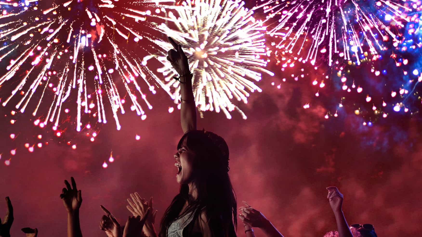 A young woman raises her arm in celebration against a backdrop of brightly coloured fireworks in the sky.
