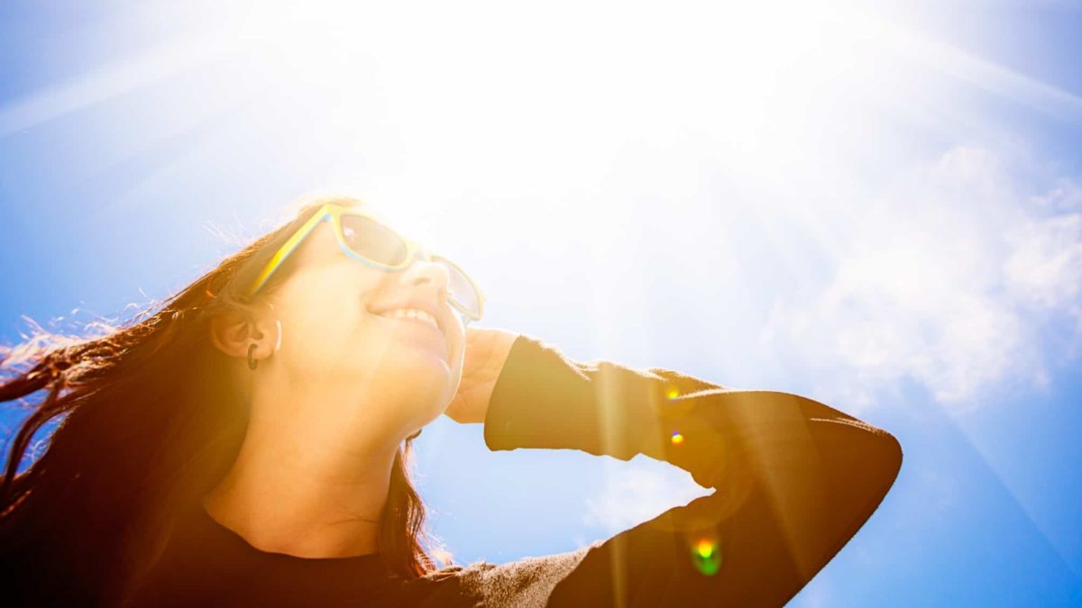 A woman wears sunglasses as she gazes up towards a bright sun with its rays extending to the far corners of the sky above her.