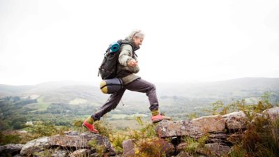 a woman in full hiking gear carrying a backpack and camping equipment on her back takes a large step between two sections of a rocky pathway in a misty outdoor setting.